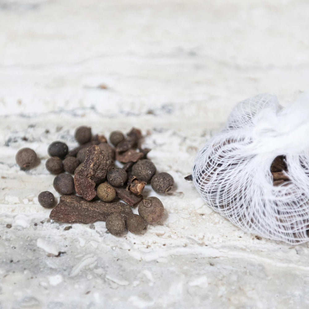 Spices including black peppercorns and star anise on a light surface with a white mesh bag.