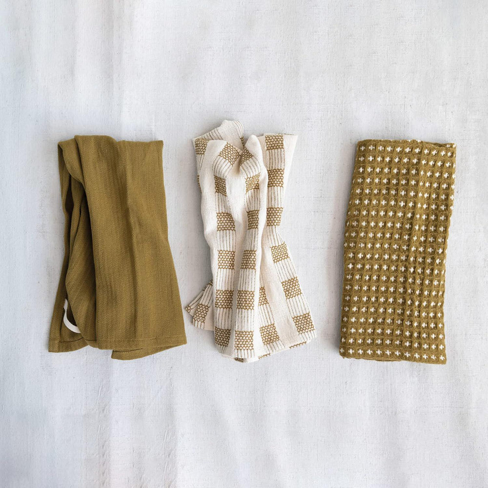 Three folded fabric swatches in green, white with brown pattern, and gold with white pattern on a light gray background.