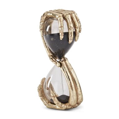 A decorative hourglass figurine featuring skeletal hands in a metallic gold finish holding a clear glass hourglass filled with dark sand, set against a plain white background.