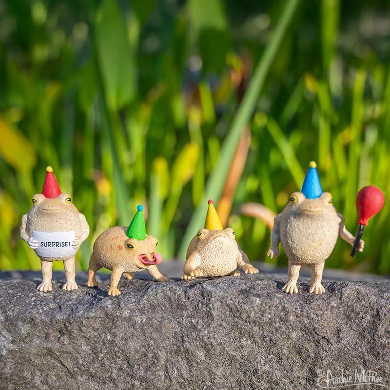 Five small frog figurines with colorful hats and a red balloon on a stone surface with a green leafy background.