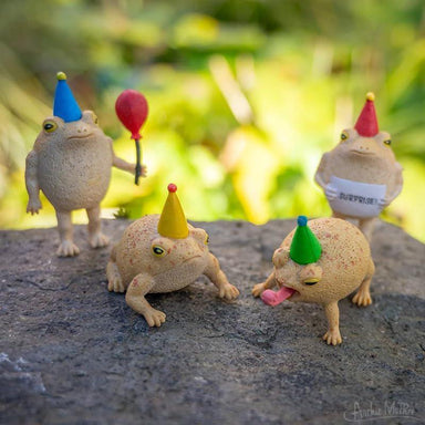 Four toy frogs with colorful hats on a stone surface with a blurred natural background