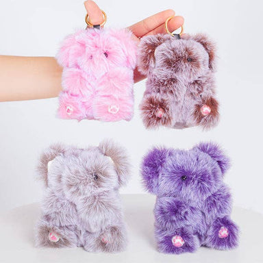 Four plush teddy bear phone cases in pink, brown, light gray, and purple, with small pink paw pads and gold key rings, shown on a clean white background with one case held by a hand.
