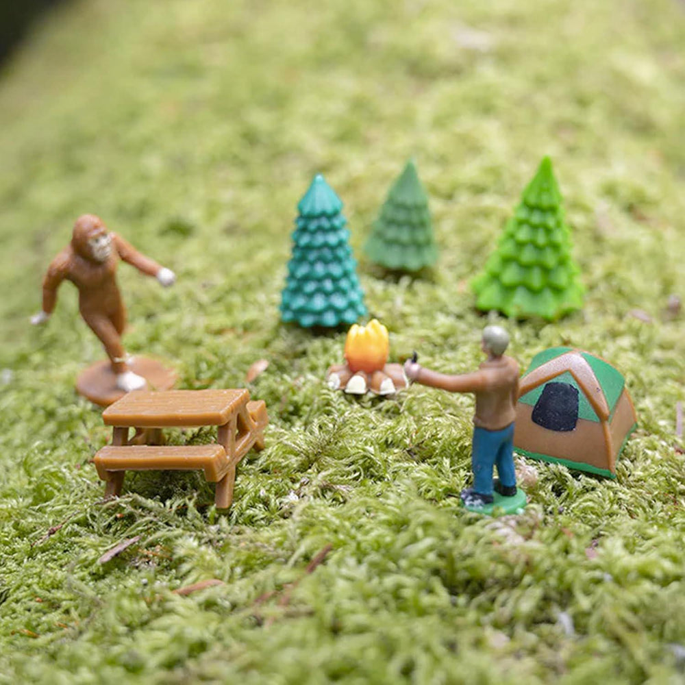 Miniature figures of a person, a tent, a picnic table, and trees on a grassy surface.