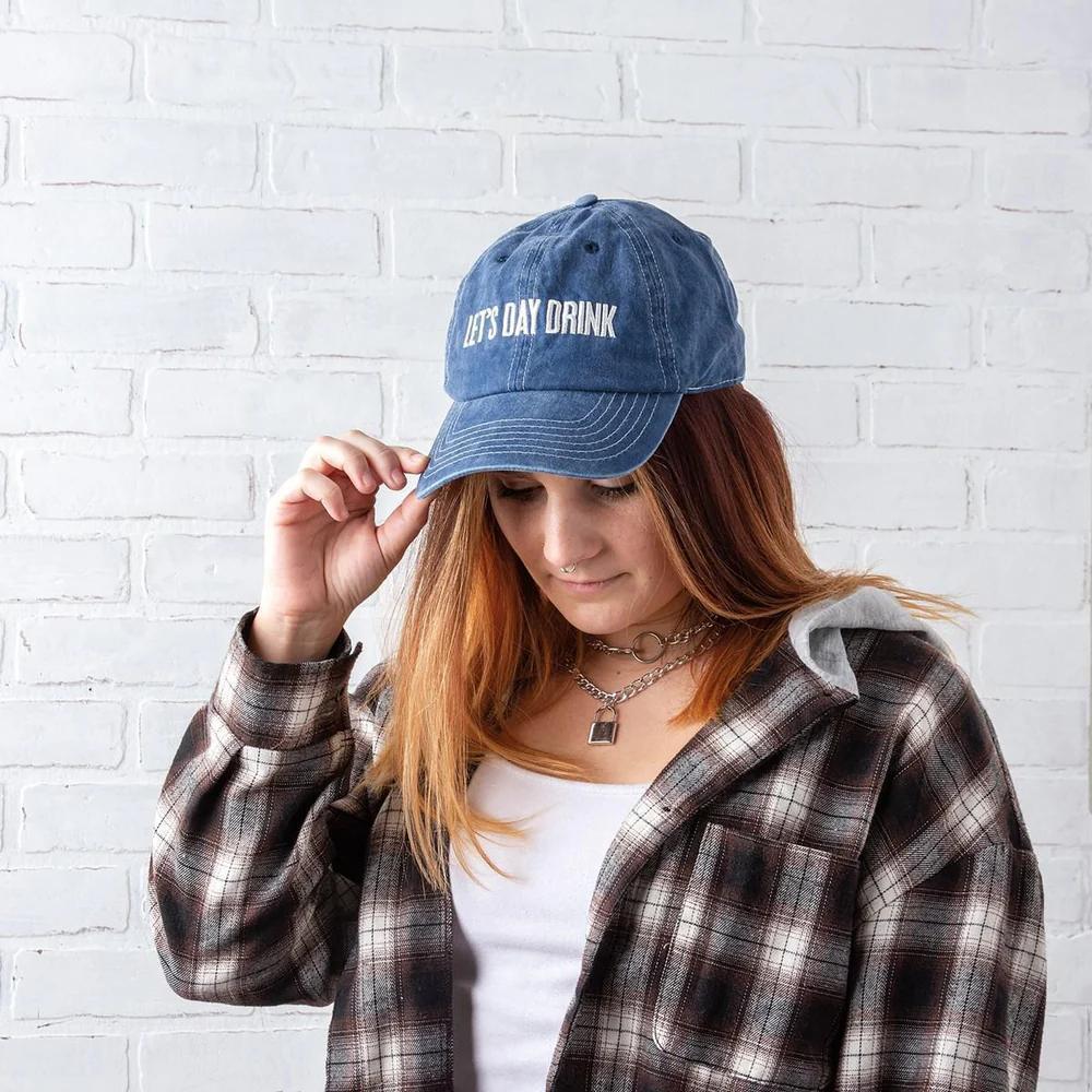 Person wearing a blue cap with 'Let's Day Drink' text against a white brick wall.