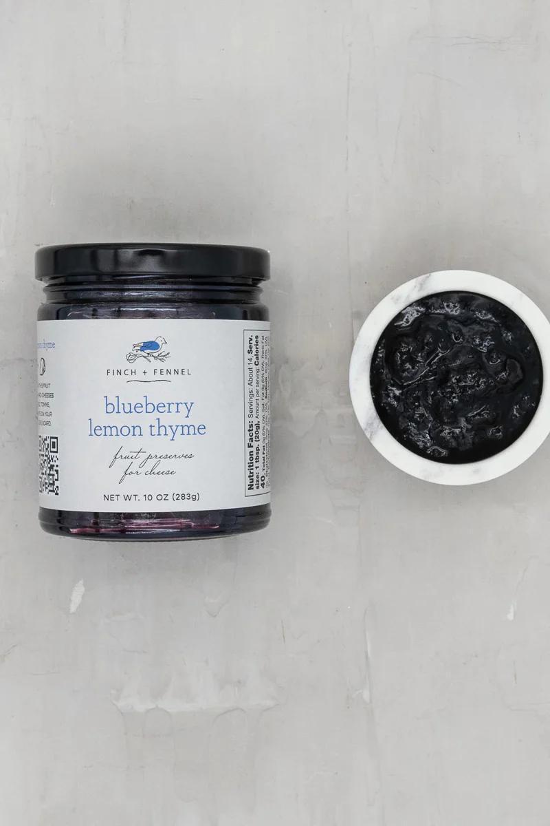 Jar of 'Blueberry Lemon Thyme' preserve with a small dish of the preserve on a gray background