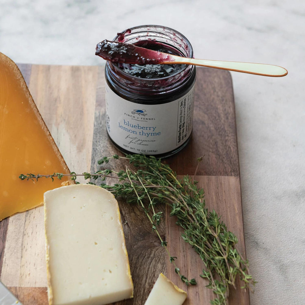 Jar of blueberry lemon thyme jam on a wooden cutting board with cheese and herbs.