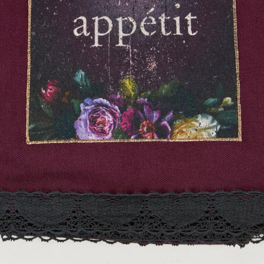 A close-up of a textile product, likely a scarf or bandana, featuring a deep burgundy fabric body, a black lace trim along the bottom edge, and a rectangular printed panel with the word 'appétit' above a floral illustration in purple, pink, green, and cream tones.