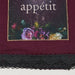 A close-up of a textile product, likely a scarf or bandana, featuring a deep burgundy fabric body, a black lace trim along the bottom edge, and a rectangular printed panel with the word 'appétit' above a floral illustration in purple, pink, green, and cream tones.