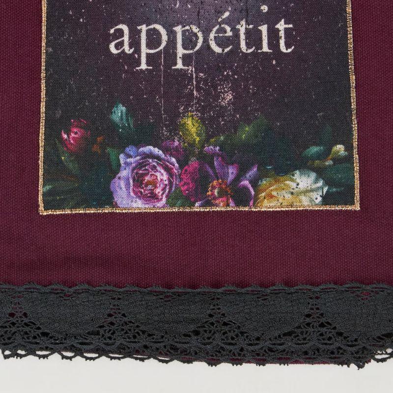 A close-up of a textile product, likely a scarf or bandana, featuring a deep burgundy fabric body, a black lace trim along the bottom edge, and a rectangular printed panel with the word 'appétit' above a floral illustration in purple, pink, green, and cream tones.