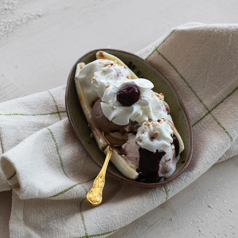 Banana split with ice cream, whipped cream, and a cherry on a plate with a gold spoon, placed on a checkered cloth.