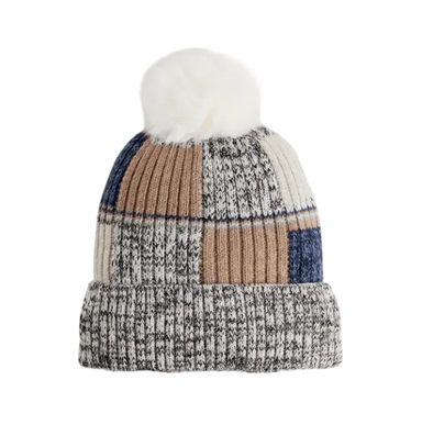 A knit beanie hat with a fluffy white pom-pom on top, featuring a patchwork design of cream, tan, and navy blue ribbed panels on a light gray background.