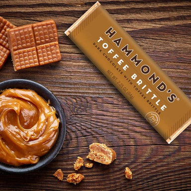 A package of Hammond's Toffee Brittle lies diagonally on a dark wooden table, surrounded by pieces of the golden-brown brittle, a small bowl of caramel sauce, and a few crumbs. The packaging is a metallic gold color with black and white text.