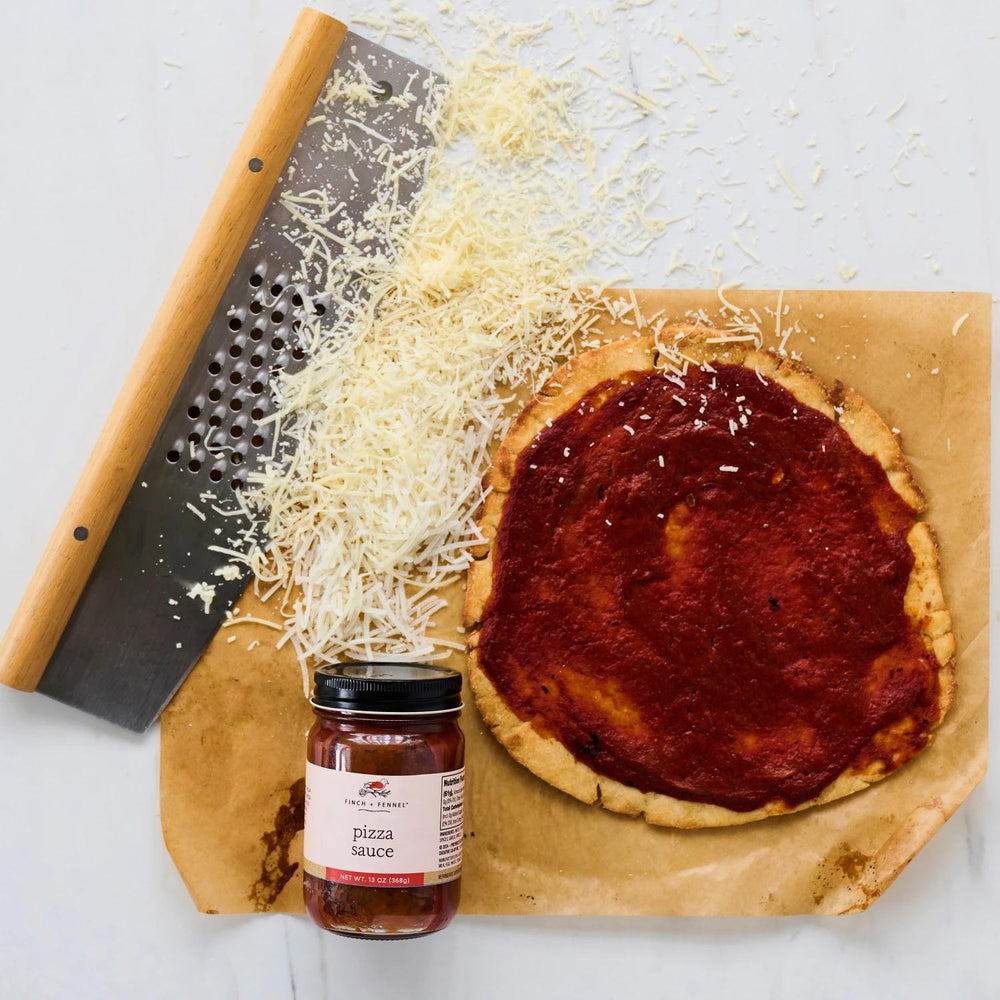 Unbaked pizza with tomato sauce on a wooden board, accompanied by a jar of pizza sauce and a grater.