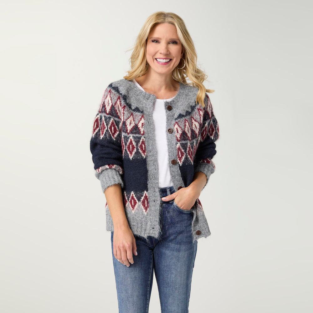 A smiling blonde woman stands against a light gray studio background wearing a gray knit cardigan with a navy and burgundy geometric pattern, a white top, and blue jeans.