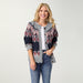 A smiling blonde woman stands against a light gray studio background wearing a gray knit cardigan with a navy and burgundy geometric pattern, a white top, and blue jeans.
