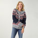 A smiling blonde woman wearing a gray knit cardigan with a navy blue yoke and red, white, and gray geometric patterns, paired with blue jeans, against a light gray studio background.