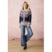 A woman models a navy blue cardigan with a gray fuzzy collar and red, white, and blue geometric patterns, paired with wide-leg blue jeans and dark blue boots, standing against a pink textured backdrop.