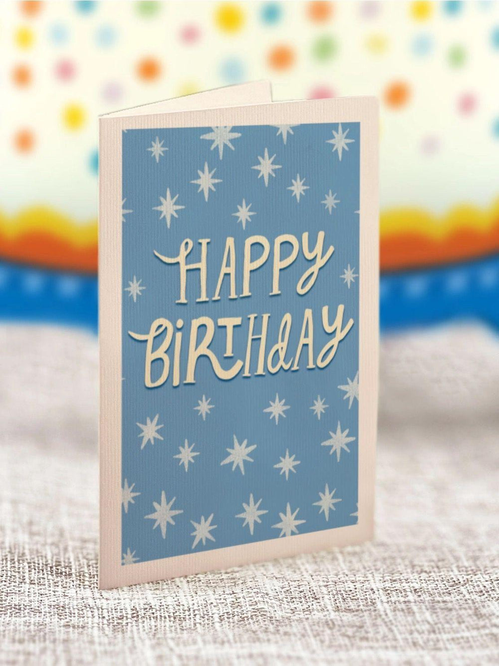 Blue birthday card with white star patterns and 'Happy Birthday' text on a textured surface with colorful blurred background.