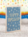 Blue birthday card with white star patterns and 'Happy Birthday' text on a textured surface with colorful blurred background.
