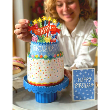 Colorful birthday cake with candles and a 'Happy Birthday' sign, held by a person.