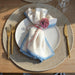 Dining table setting with a white plate, gold fork and knife, and a folded napkin with a decorative flower.