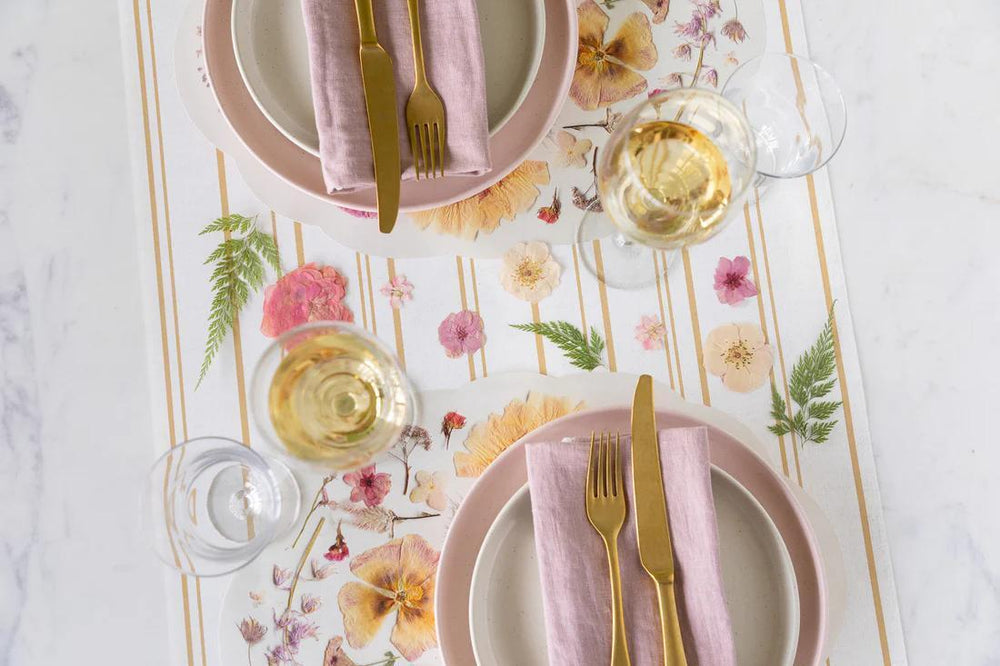 Elegant table setting with floral placemat, pink plates, gold cutlery, and white wine glasses.