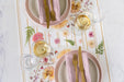 Elegant table setting with floral placemat, pink plates, gold cutlery, and white wine glasses.