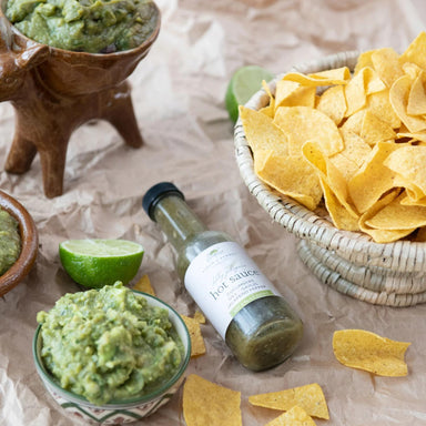 Bottle of hot sauce with bowls of guacamole and chips on a textured surface