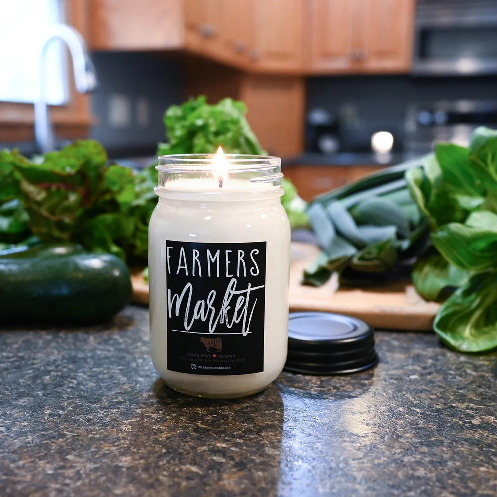 Candle labeled 'Farmers Market' on a kitchen counter with produce in the background