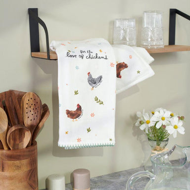 Kitchen towel with chicken design hanging on a wall next to wooden utensils and a shelf.