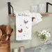 Kitchen towel with chicken design hanging on a wall next to wooden utensils and a shelf.
