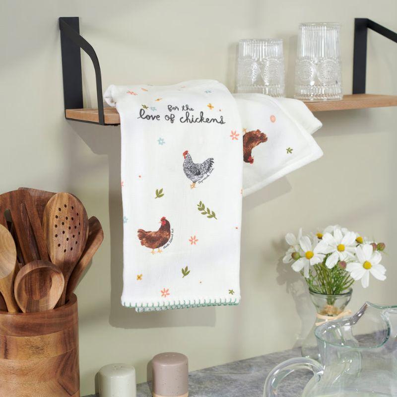 Kitchen towel with chicken design hanging on a wall next to wooden utensils and a shelf.