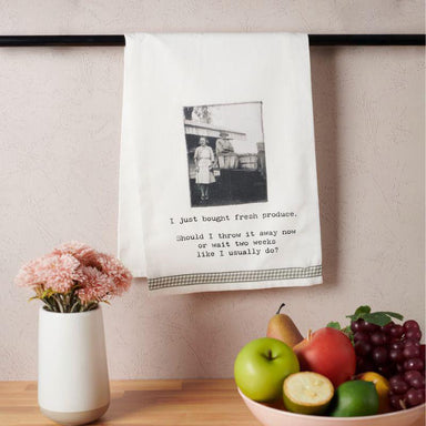 Kitchen towel with a humorous print hanging on a rack, next to a bowl of fruit and a vase with flowers.