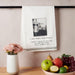 Kitchen towel with a humorous print hanging on a rack, next to a bowl of fruit and a vase with flowers.