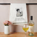 Towel with a printed design and text, wine bottle, glass, and flowers on a wooden surface.