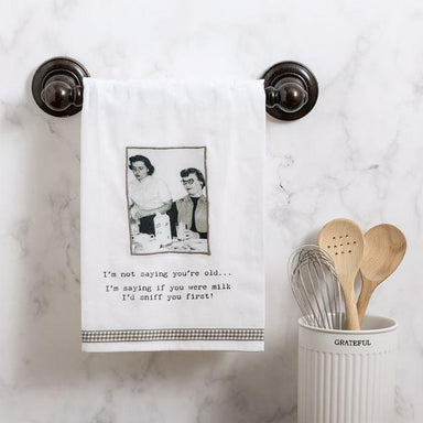 Kitchen towel with humorous text and photo hanging on a marble wall, next to wooden spoons and a whisk in a container.