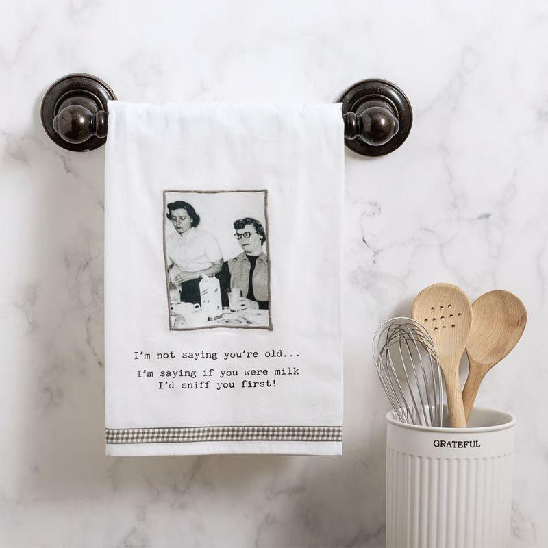 Kitchen towel with humorous text and photo hanging on a marble wall, next to wooden spoons and a whisk in a container.