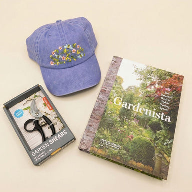 Blue cap with floral design, garden shears packaging, and 'Gardenista' book on a beige background