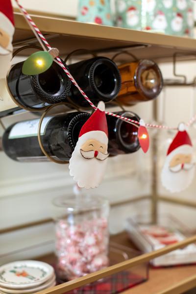 Decorative wine bottle holder with Santa Claus figures and string lights on a shelf.