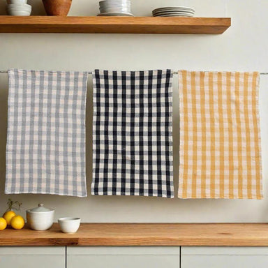 Three checkered towels in gray, black, and yellow hanging on a rack with a kitchen setting in the background.