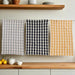 Three checkered towels in gray, black, and yellow hanging on a rack with a kitchen setting in the background.