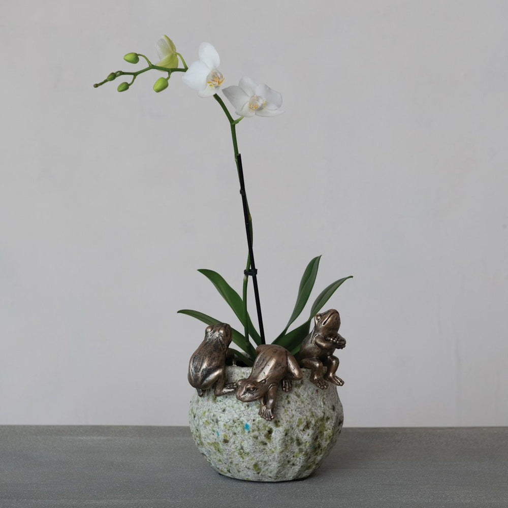 Decorative vase with three frog sculptures and a white orchid on a gray surface.