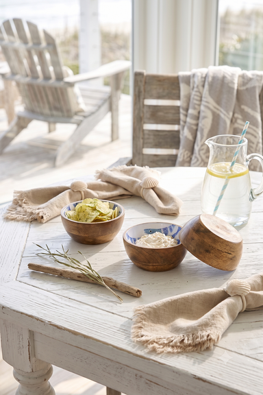 A styled tabletop scene with two small wooden bowls containing dips, a glass pitcher of water with a lemon slice and straw, beige napkins, and a rustic white table in a bright indoor setting with chairs and a window in the background.