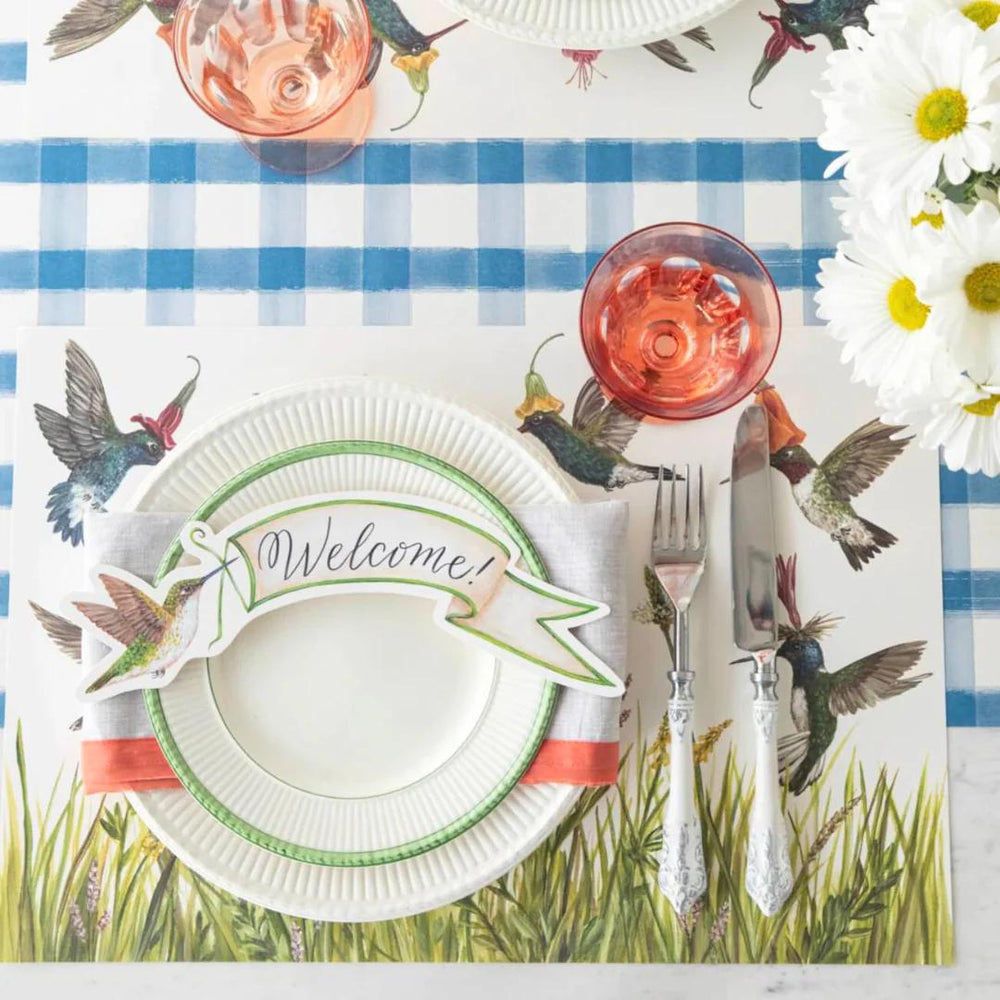 Table setting with 'Welcome' plate, red glass, and hummingbird-themed placemat.