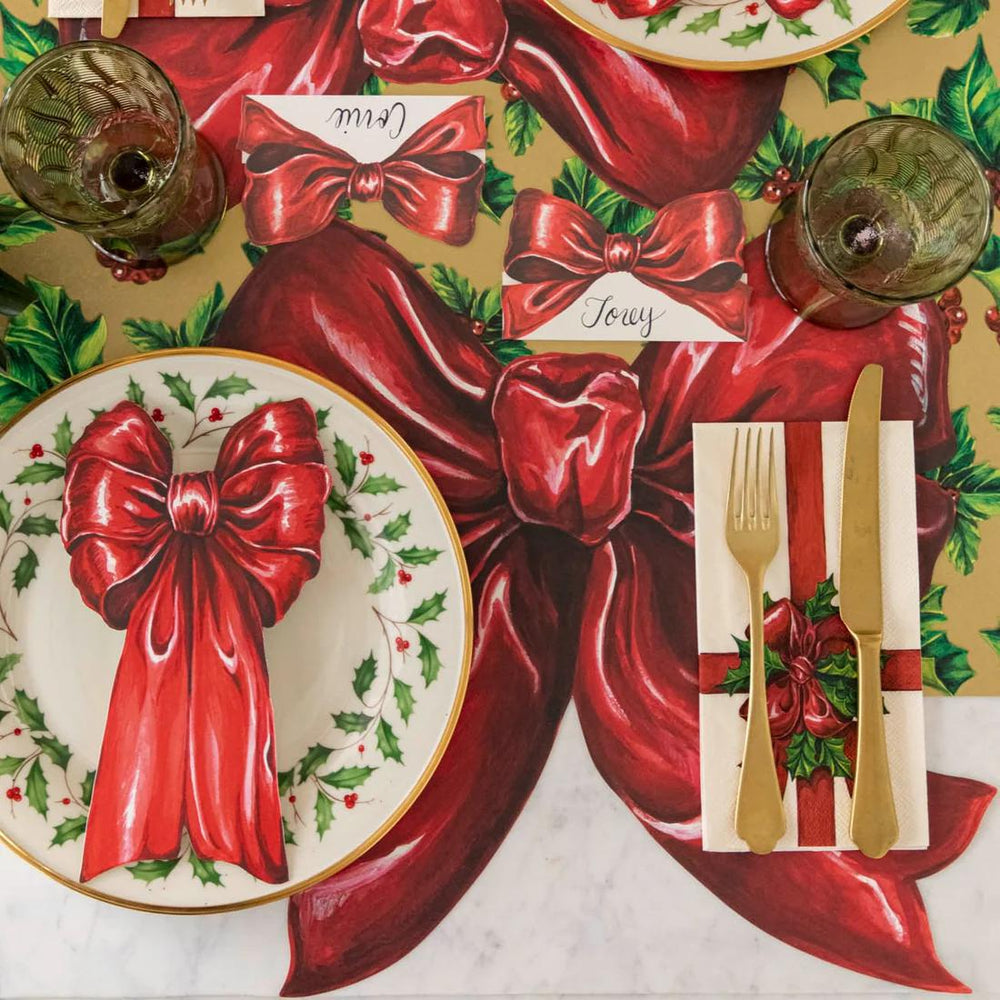 Christmas-themed table setting with red bows, gold cutlery, and holly leaves on a decorative paper background.