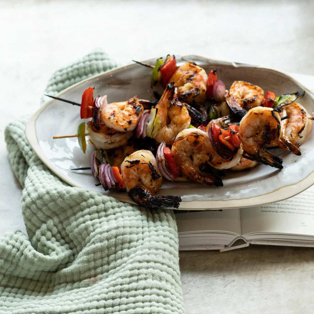 Grilled shrimp skewers on a plate with a green towel and open book in the background