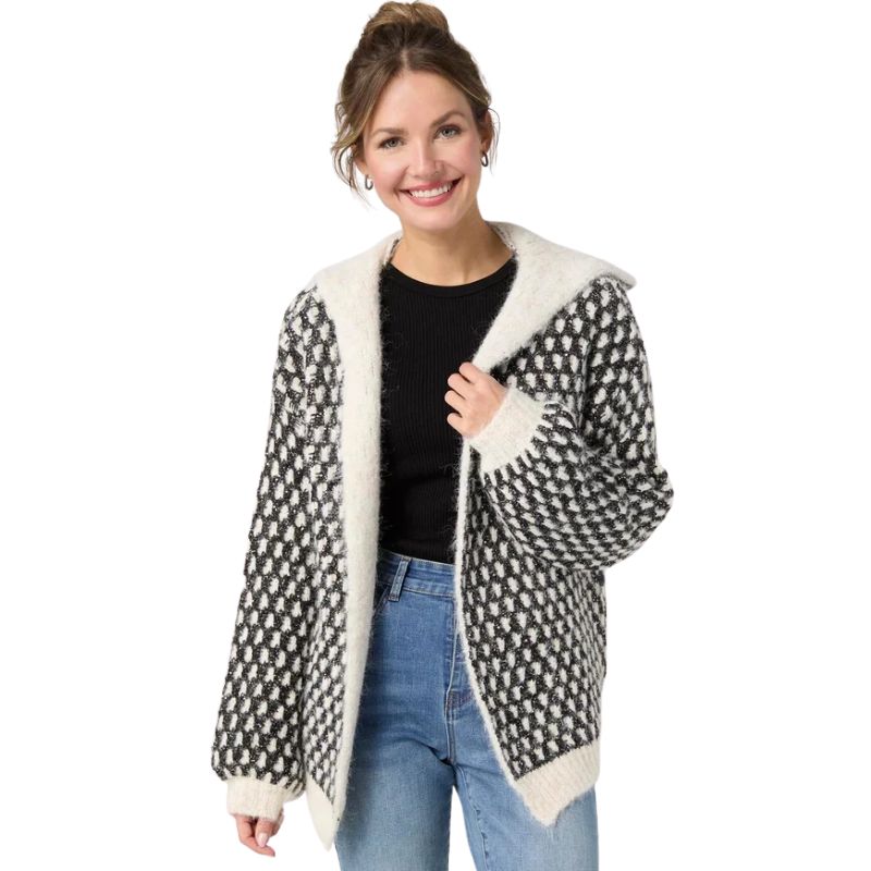 A woman modeling a black and white geometric patterned cardigan with a cream-colored fuzzy sherpa-lined hood and cuffs, worn over a black top and blue jeans against a plain white background.
