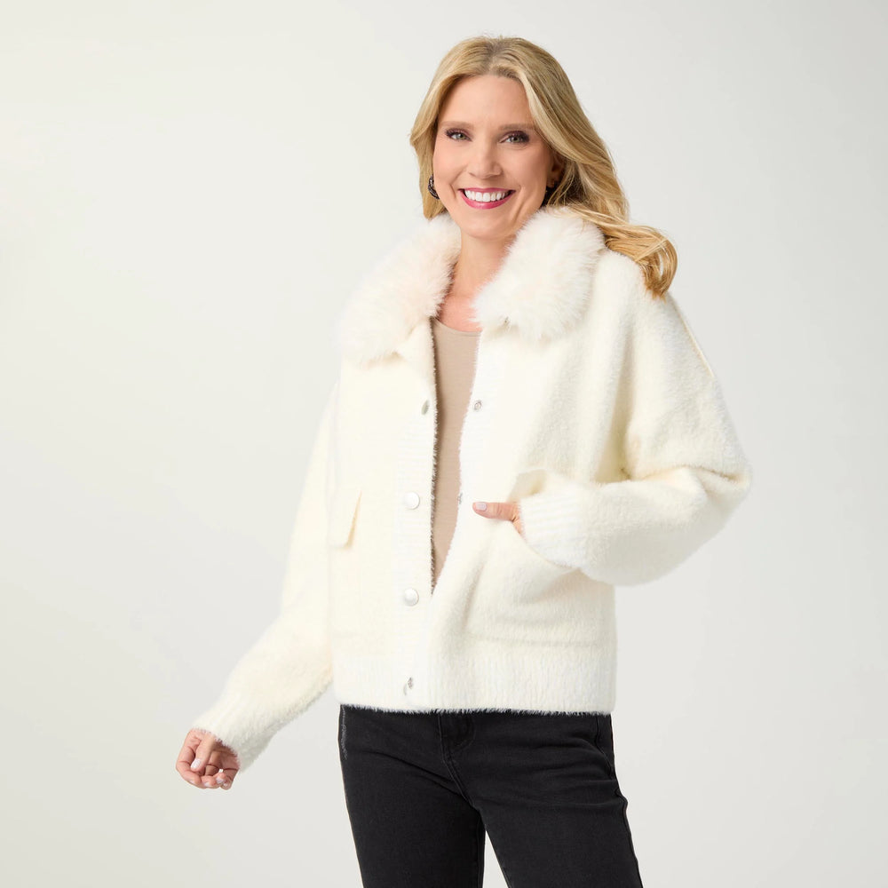 A smiling blonde woman wearing a cream-colored fuzzy button-front cardigan with a large fluffy collar, paired with a beige top and black pants, photographed against a light gray studio background.