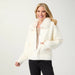A smiling blonde woman wearing a cream-colored fuzzy button-front cardigan with a large fluffy collar, paired with a beige top and black pants, photographed against a light gray studio background.