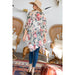 A woman wearing a bohemian-style floral kimono top, blue jeans, and a straw hat stands in a decorated room with wicker chairs and a rug, photographed from behind.